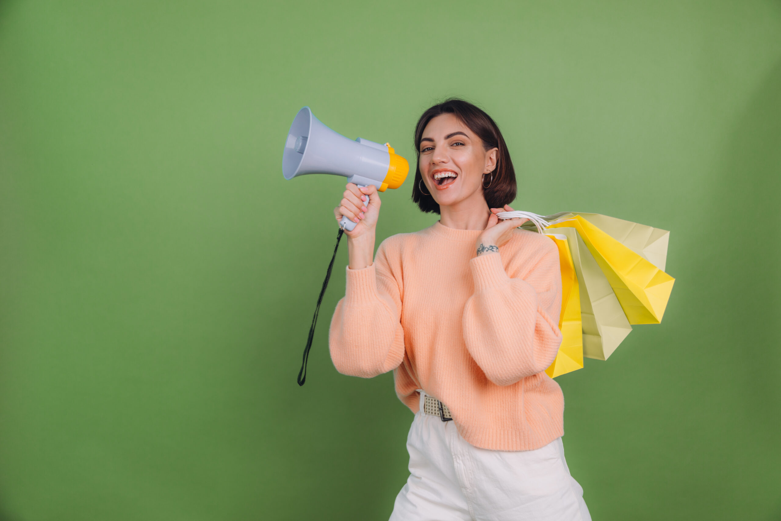 Young woman in casual peach sweater isolated on green olive color background shout in megaphone holding shopping bags, announces discounts sale promotion